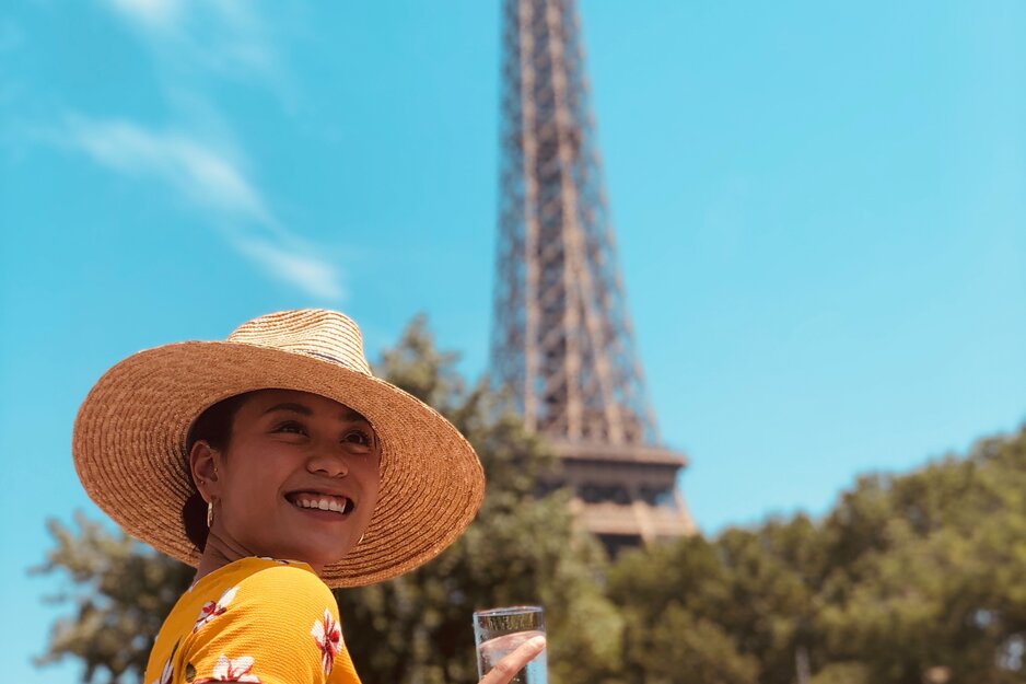 Junge Frau im gelben Kleid und Sonnenhut mit Drink vor Eiffelturm | © Unsplash