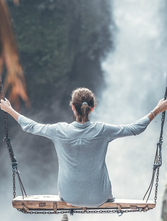 Urlauberin sitzt auf Holzschaukel vor Wasserfall | © Pexels