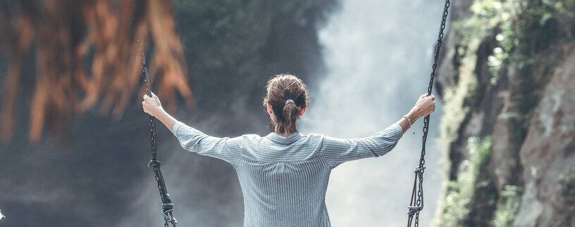 Urlauberin sitzt auf Holzschaukel vor Wasserfall | © Pexels