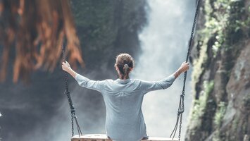 Urlauberin sitzt auf Holzschaukel vor Wasserfall | © Pexels