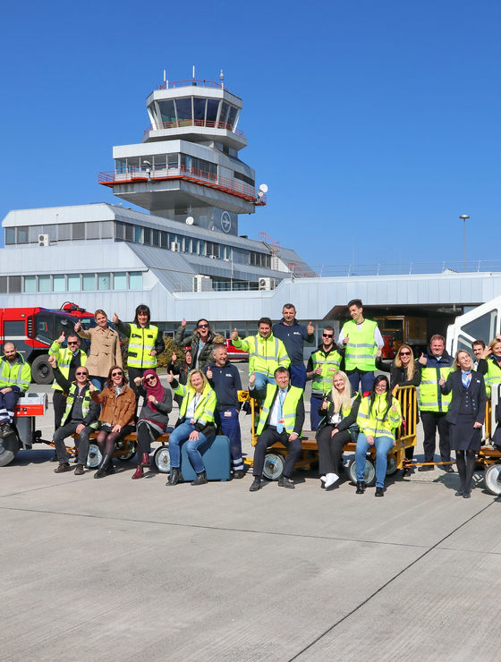 Flughafenmitarbeiter sitzen winkend auf Gepäckswagerl am Vorfeld vor dem Tower | © Linz Airport