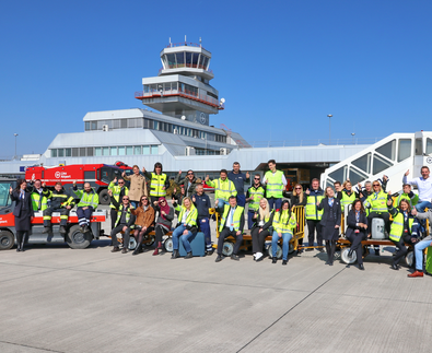 Flughafenmitarbeiter sitzen winkend auf Gepäckswagerl am Vorfeld vor dem Tower | © Linz Airport