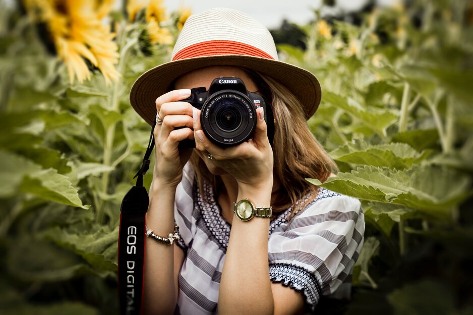 Junge Frau mit Sonnenhut mit Kamera vor dem Gesicht in einem Sonnenblumenfeld | © Pexels