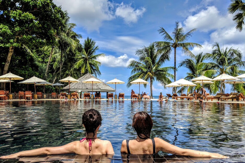 Zwei Frauen schauen vom Pool aus auf Palmen und Strand | © Unsplash