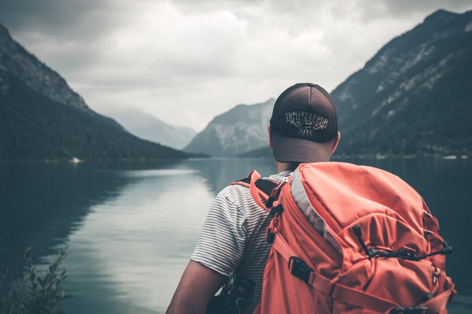 Junger Wanderer mit Kappe und Rucksack blickt auf Bergsee | © Philipp Kammerer Unsplash