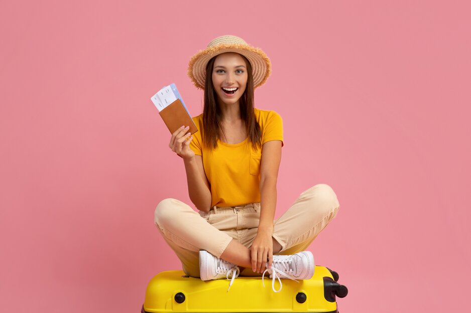 Fröhliche junge Frau mit Sonnenhut und Flugtickets sitz auf einem gelben Koffer